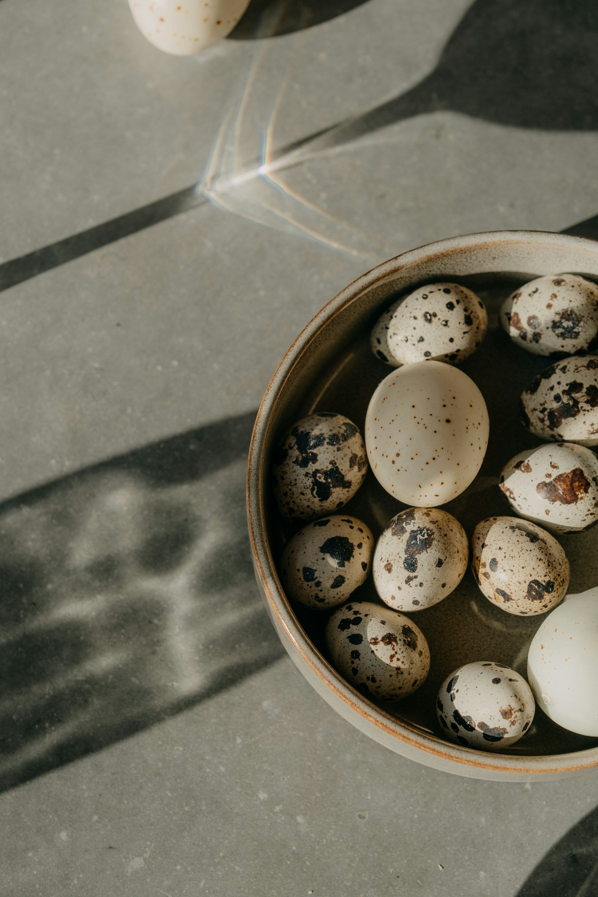 A bowl of quail eggs on a table next to a shadow of a glass.