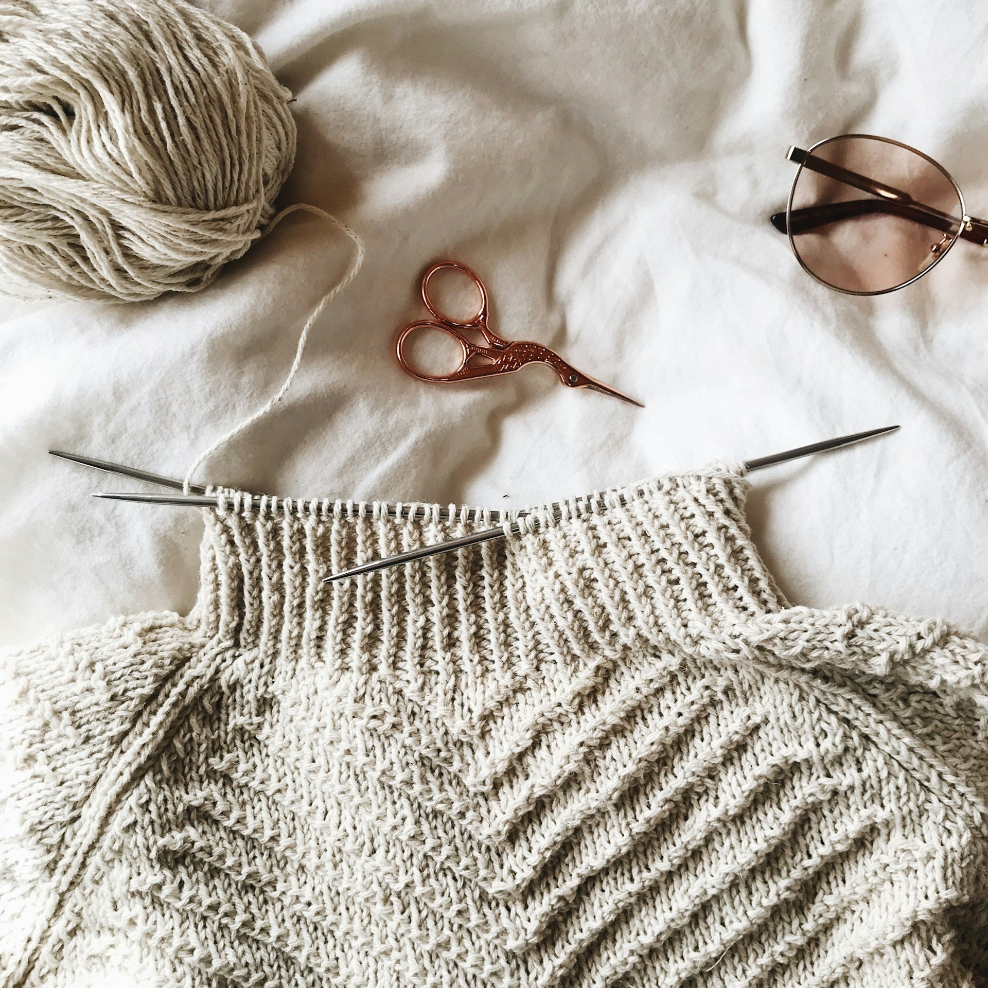 A sweater, knitting needles, and a pair of sunglasses on a bed.
