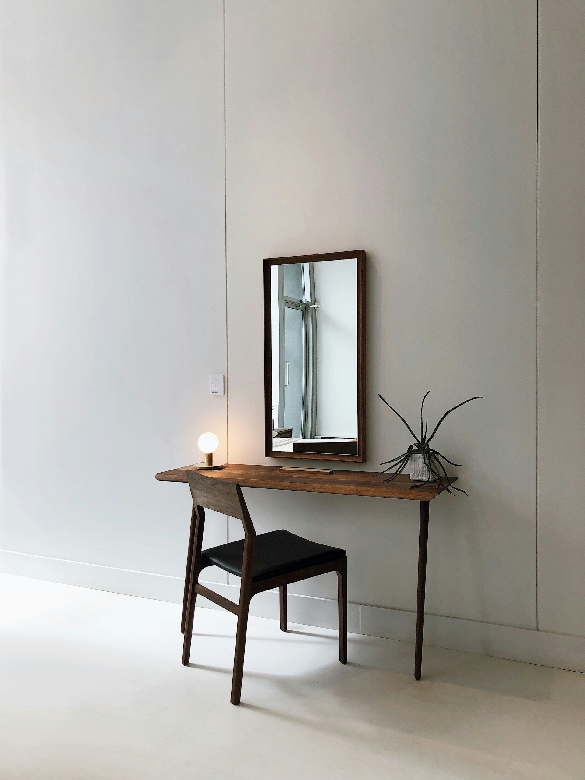 A wooden desk with a mirror and chair in front of a white wall.